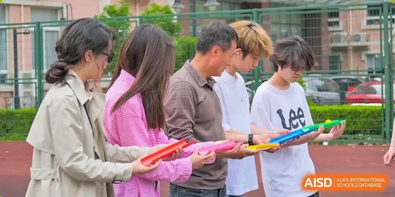 A group of students and an older person stand closely together outdoors, receiving colorful cylindrical objects during a planned group activity or event, at Cambridge International School Litai College. SISU.