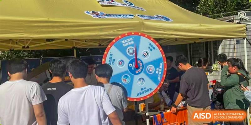 Students participate in an event, gathering around a large blue prize wheel under a bright yellow tent at Cambridge International School Litai College. SISU.