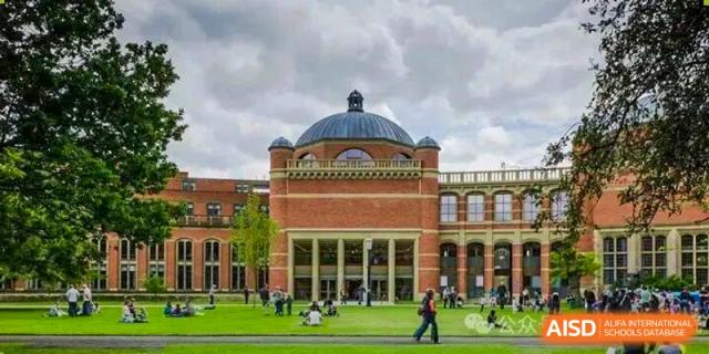 A large, classic red brick school building featuring a prominent domed roof and arches stands behind a green lawn at Cambridge International School Litai College. SISU.