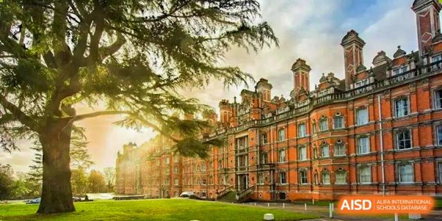 The facade of an imposing, traditional red brick college building with multiple chimneys is seen through sunlit trees on the campus lawn at Cambridge International School Litai College. SISU.