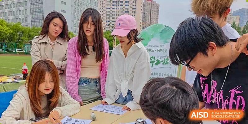 Two young women, one in a pink cap, look down intently at papers on a table during an outdoor student sign-up or informational event at Cambridge International School Litai College. SISU.
