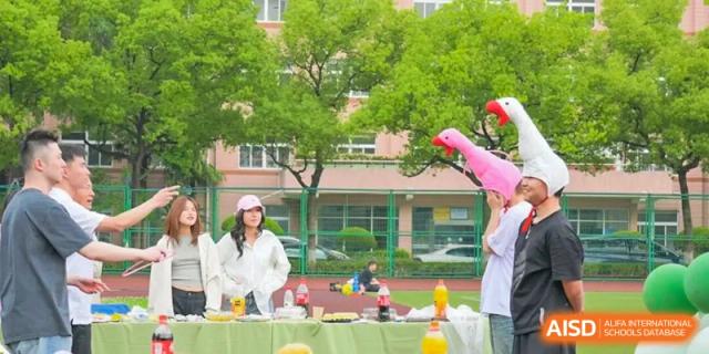 A group of students is gathered around a table with a yellow cloth on a grassy area during an outdoor function near the main school building at Cambridge International School Litai College. SISU.