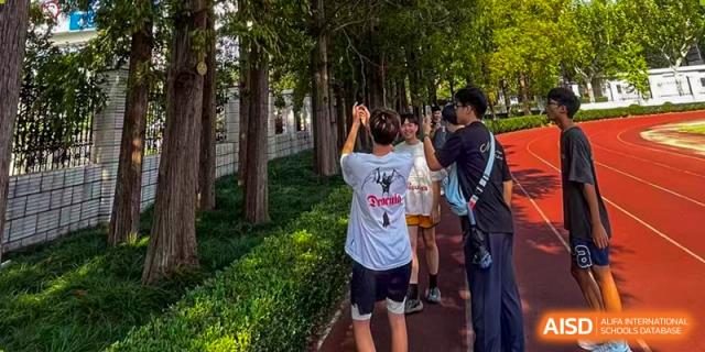 Students are seen outdoors, with one student actively taking a photo, along a path lined with trees at Cambridge International School Litai College. SISU.