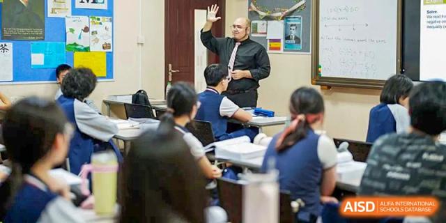 A teacher in a black shirt raises his hand while lecturing a classroom of attentive students in uniform at the Shenzhen Academy of International Education.