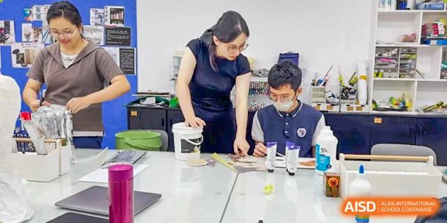 A teacher assists a student with an art project involving paint tubes and materials on a large work table at the Shenzhen Academy of International Education.