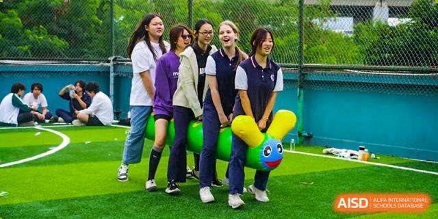 Students participate in a cooperative race using a large, green inflatable caterpillar on a field at Shenzhen Academy of International Education.