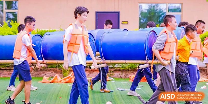 Students wearing life jackets practice teamwork by carrying large blue barrels for a creative building challenge at Shenzhen Academy of International Education.