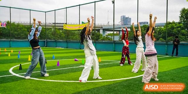 Students in white and red athletic wear practicing a synchronized dance routine on a bright green outdoor sports field at Shenzhen Academy of International Education.
