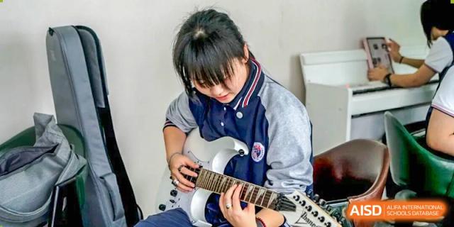 A student focuses intently while playing a sleek silver electric guitar during a music practice or performance session at Shenzhen Academy of International Education.