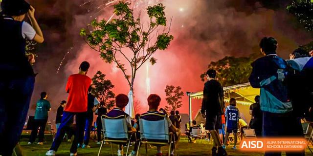 Students gather outdoors at night to witness a spectacular rocket launch illuminating the sky behind a leafy tree at Shenzhen Academy of International Education.