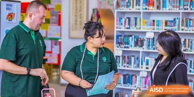 A staff member wearing a green polo shirt holds documents while standing in front of library bookshelves at Shen Wai International School.