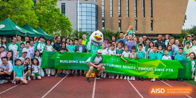 Students and staff pose behind a green banner reading "Building Bridges, Nurturing Minds" alongside a school mascot on a running track at Shen Wai International School.