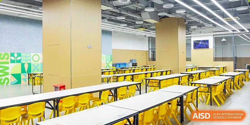 A bright and modern school cafeteria featuring long white tables and vibrant yellow chairs, organized and ready for student dining at Shen Wai International School.