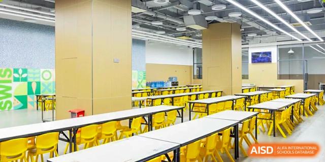 A bright and modern school cafeteria featuring long white tables and vibrant yellow chairs, organized and ready for student dining at Shen Wai International School.