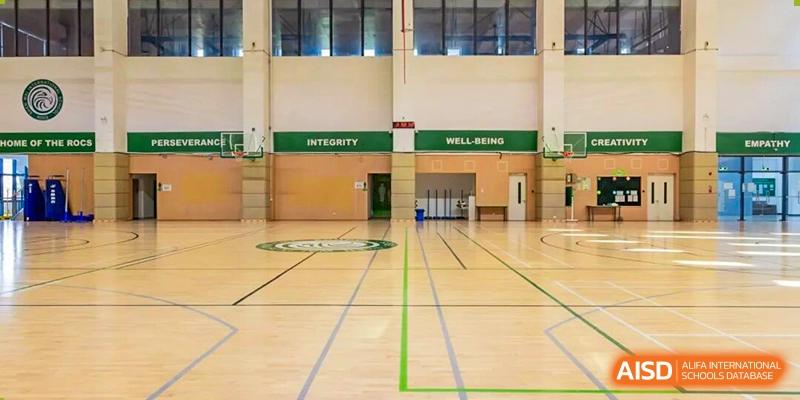 An empty indoor school gymnasium featuring polished wooden floors and green banners on the wall labeled "Integrity" and "Well-Being" at Shen Wai International School.