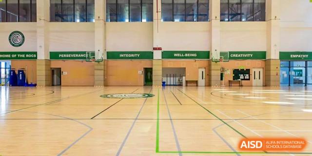 An empty indoor school gymnasium featuring polished wooden floors and green banners on the wall labeled "Integrity" and "Well-Being" at Shen Wai International School.