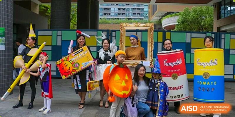 Teachers and students dressed in creative costumes, including a pumpkin and a framed painting, pose together for a festive outdoor celebration at Shen Wai International School.