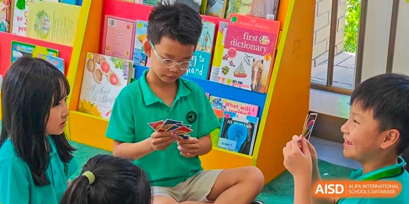 A young male student in a green polo shirt sits on the floor and focuses on a hand of playing cards in front of a colorful book display at Shen Wai International School.