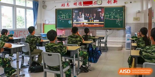 Students in camouflage uniforms sit at their desks while watching an educational broadcast on a large screen at Zhongshan Sanxin School International Department.