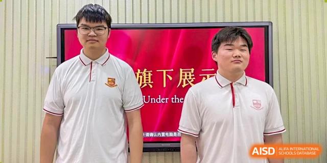 Two students in white polo shirts stand in front of a bright red digital display screen during a formal school presentation at Zhongshan Sanxin School International Department.