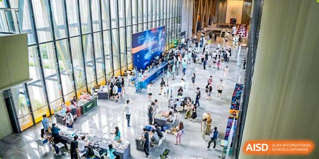 A high-angle view of a bustling, modern glass-walled hall filled with people visiting various information booths and displays at Zhongshan Sanxin School International Department.