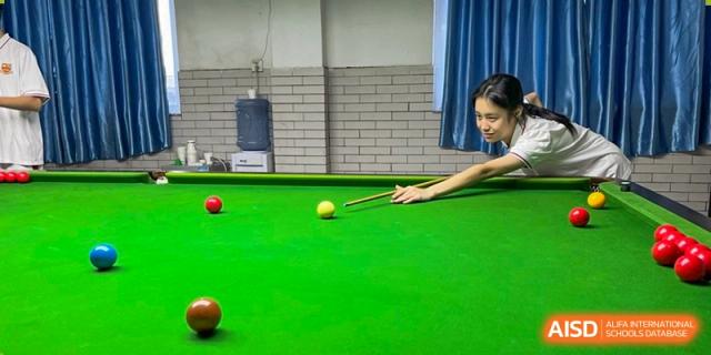 A close-up of a student focused on a billiards table, carefully aiming a pool cue at a yellow ball during an extracurricular activity at Zhongshan Sanxin School International Department.