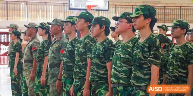 A row of male students wearing green camouflage uniforms and matching caps stand at attention during a military training event at Zhongshan Sanxin School International Department.