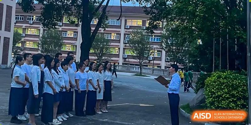 A group of students in white shirts stands at attention while an instructor with a clipboard addresses them outside a campus building at Zhongshan Sanxin School International Department.