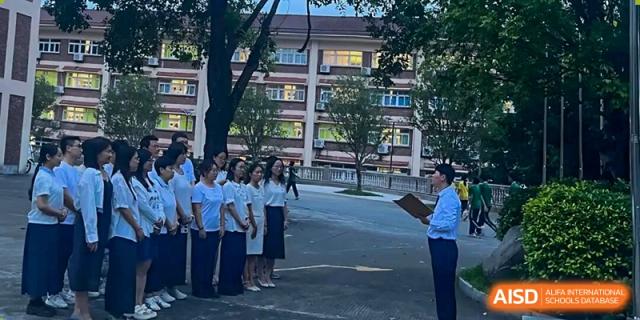 A group of students in white shirts stands at attention while an instructor with a clipboard addresses them outside a campus building at Zhongshan Sanxin School International Department.