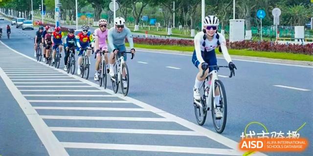 A group of cyclists wearing helmets and athletic gear ride in a line along a paved road during an outdoor fitness excursion at Zhongshan Sanxin School International Department.