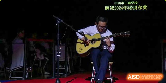 A student sits on a stage and performs a solo acoustic guitar piece during a talent showcase or musical event at Zhongshan Sanxin School International Department.