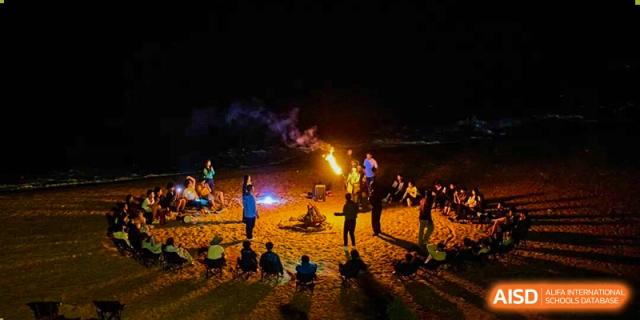 Students gather around a glowing bonfire on a beach at night for a social gathering and team-building event at Zhongshan Sanxin School International Department.