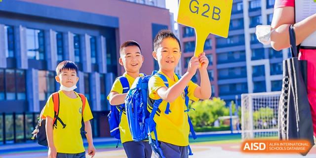 Two young elementary students in bright yellow shirts walk together on the sunny school campus while one student holds up a yellow "G2B" class sign at Beijing Chaoyang KaiWen Academy.
