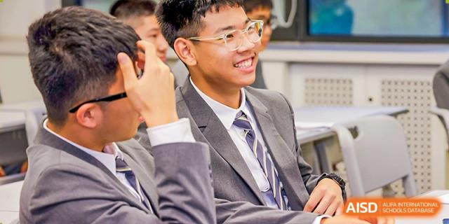 A male student wearing a formal grey suit and striped tie smiles broadly while participating in a classroom discussion with his peers and teacher at Beijing Chaoyang KaiWen Academy.