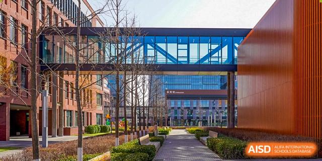 A scenic view of the modern campus architecture shows a sleek glass skybridge connecting two brick academic buildings under a clear blue sky at Beijing Chaoyang KaiWen Academy.