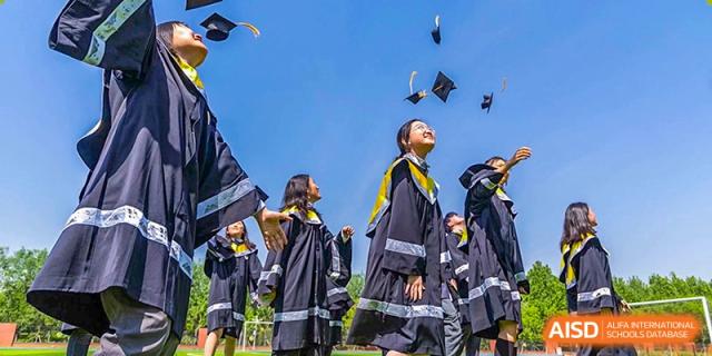 Graduates in black and yellow robes celebrate their academic success by tossing their mortarboard caps high into the air on a sunny day at Beijing Chaoyang KaiWen Academy.