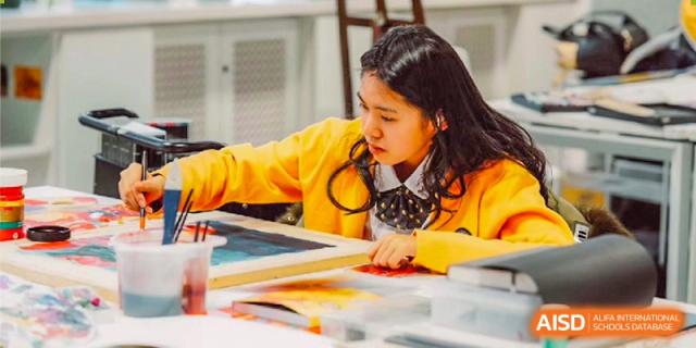A focused female student in a yellow sweater works diligently on a detailed painting in an art studio filled with creative supplies at Beijing Chaoyang KaiWen Academy.