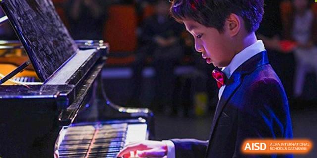 A young male student in a formal suit and red bowtie concentrates while performing a classical piece on a grand piano during a recital at Beijing Chaoyang KaiWen Academy.