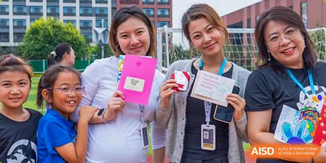 Two smiling staff members proudly display handmade cards and crafts received during a heartwarming teacher appreciation event at Beijing Chaoyang KaiWen Academy.