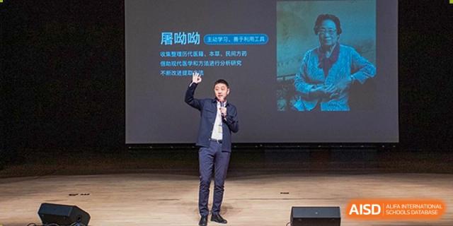 A professional speaker stands on a stage and gestures toward a presentation slide featuring a famous scientist during an assembly at Beijing Chaoyang KaiWen Academy.