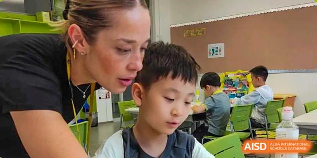 A teacher observes a young boy closely as he works on a detailed task at his desk, providing personalized academic support in a bright classroom at Beijing Chaoyang KaiWen Academy.