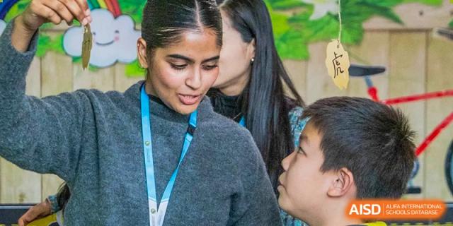 A female teacher with a blue lanyard leans in to have a focused one-on-one conversation with a young student during an interactive classroom session at Beijing Chaoyang KaiWen Academy.