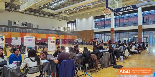 Groups of parents and teachers sit at round tables in a large gymnasium for a collaborative workshop or community networking event at Beijing Chaoyang KaiWen Academy.