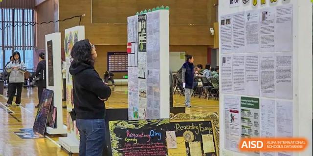 A student stands and carefully reviews a detailed research poster displayed on a board during a formal academic science fair held in the gymnasium at Beijing Chaoyang KaiWen Academy.