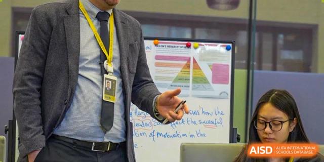 A teacher in a grey blazer points toward a whiteboard displaying Maslow's hierarchy of needs during a social studies or psychology lecture at Beijing Chaoyang KaiWen Academy.