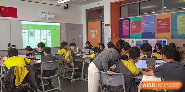 A teacher leans over a desk to assist two students who are working together on a laptop during a technology-integrated classroom activity at Beijing Chaoyang KaiWen Academy.