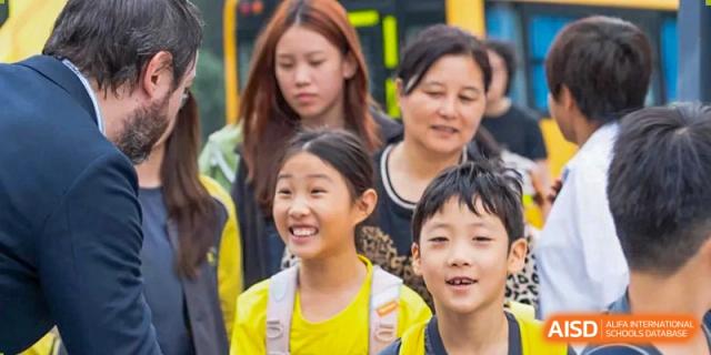 A teacher and several young students in yellow and white shirts smile together during a fun and social outdoor gathering on the school grounds at Beijing Chaoyang KaiWen Academy.
