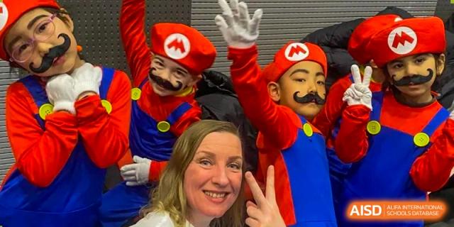 A teacher smiles for a photo with two young students who are playfully dressed up in red and blue Mario costumes during a fun school event at Beijing Chaoyang KaiWen Academy.