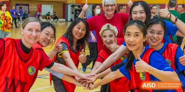 A diverse group of female staff and faculty members in colorful team shirts smile and join their hands together in a circle of unity during a sports event at Beijing Chaoyang KaiWen Academy.