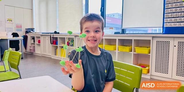 A young boy smiles proudly while holding up a geometric structure he built using green clay and sticks during a hands-on STEAM activity in his classroom at Beijing Chaoyang KaiWen Academy.